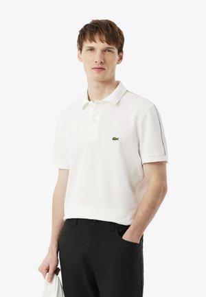 Camiseta polo blanca con un logo verde de cocodrilo, mangas cortas y cuello clásico; combinada con pantalones negros y sostenida con un paño blanco.