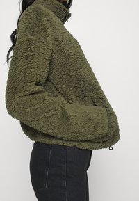 Chaqueta de forro polar color verde oliva con cuello alto, superficie texturizada y bolsillos laterales, diseñada para proporcionar calidez y comodidad.