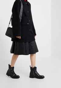 Abrigo de lana negro con grandes botones, combinado con una falda de cuero negro con pliegues y botas negras de tobillo con cordones y cremalleras laterales.
