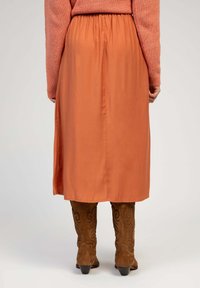 Jupe midi couleur rouille avec une taille élastique, tissu lisse et silhouette fluide, associée à des bottes de cowboy marron ornées de motifs raffinés.