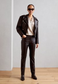 Veste de moto en cuir noir avec zippers, blouse de couleur claire et pantalon en cuir noir ajusté. Les accessoires incluent des lunettes de soleil et des boucles d'oreilles créoles.