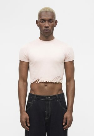 Muž s krátkymi zosvetlenými vlasmi v priliehavom svetloružovom krátkom tričku a čiernych vysokých nohaviciach na jednoduchom bielom pozadí.