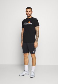 Black Ellesse t-shirt with gray logo, paired with dark shorts featuring white side stripes. Model wears white Nike shoes and socks.