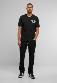 Zwarte katoenen t-shirt met korte mouwen, met een wit grafisch logo op de linkerborst. Gecombineerd met zwarte jeans en zwarte sneakers.