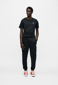 Μαύρο βαμβακερό μπλουζάκι με λογότυπο στο στήθος, συνδυασμένο με μαύρη φόρμα με πλαϊνές τσέπες. Το σύνολο ολοκληρώνουν φωτεινά κόκκινα αθλητικά παπούτσια.