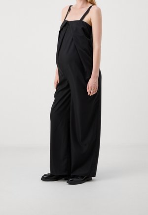 Kvinde iført løs sort ærmeløs jumpsuit med vide ben og sorte sko, stående mod en ensartet lys baggrund.