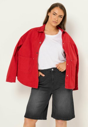 Frau trägt übergroße rote Jacke, weißes T-Shirt und lockere schwarze Denim-Shorts, steht mit einer Hand in der Tasche.