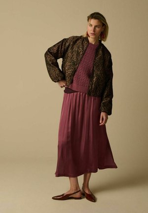 Femme portant une veste à imprimé léopard marron, un pull texturé bordeaux, une longue jupe mauve, et des ballerines marron à brides, debout devant un fond beige.
