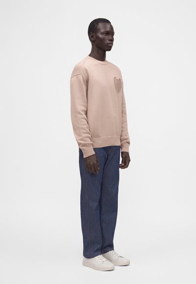 Homme debout portant un pull beige en laine avec un motif cœur sur la poitrine, un jean bleu et des baskets blanches sur un fond uni.