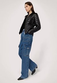 Černá péřová bunda s kapucí, zapínáním na zip a texturovaným povrchem, doplněná volnými modrými džíny typu cargo s velkými kapsami a knoflíky.