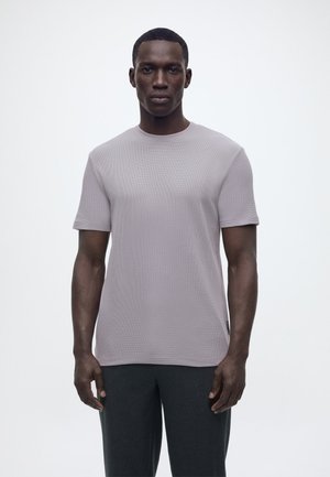 Vyras stovintis atsisukęs į priekį, dėvintis šviesiai pilką trumpomis rankovėmis tekstūruotą marškinėlį ir tamsias kelnes, stovintis priešais paprastą baltą foną.