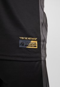 Juodi sportiniai šortai su lygiu paviršiumi, su auksiniu ir mėlynu logotipo pleistru, kuriame parašyta "MOTYVUOTIEMS" šone.