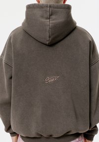 Mikina s kapucňou v tmavo šedých farbách z bavlnenej zmesi, so predným vreckom a vyšitým logom na chrbte. Mäkká textúra a oversized strih.