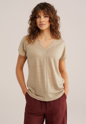 Mujer con cabello rizado que lleva una camiseta de manga corta color beige con cuello en V y pantalones burdeos, de pie con las manos en los bolsillos sobre un fondo liso.