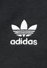 Juodas tekstilės audinys su balta siuvinėta „adidas“ logotipu, kuriame yra trys lapų formos ir trys horizontalios juostos virš prekės ženklo pavadinimo.