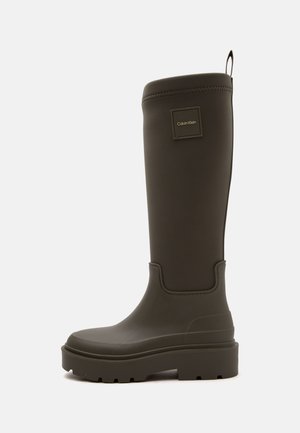 Botte de pluie haute en caoutchouc brun foncé avec semelle épaisse et écusson logo Calvin Klein près du haut sur un fond clair uni.