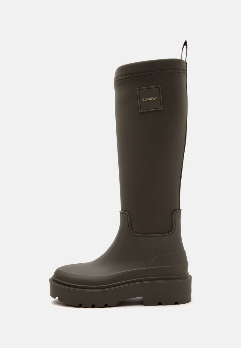 Botte de pluie haute en caoutchouc brun foncé avec semelle épaisse et écusson logo Calvin Klein près du haut sur un fond clair uni.