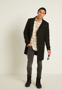 Casaco longo preto sobre uma camisa de xadrez bege, jeans slim-fit cinza e botas pretas com atacadores. Segurando óculos de sol azuis, de pé sobre um fundo claro.