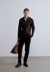 Rudas megztinis su užtrauktuku, juodos kelnės, juodi batai, rankoje nešiota ruda odinė rankinė. Minimalistinis dizainas, lygi tekstūra, neutralios spalvos.