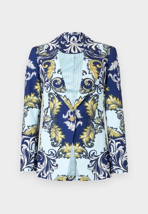 Nainen pukeutuneena laivaston siniseen bleiseriin ja siihen sopiviin shortseihin, joissa on valkoisia ja kullanvärisiä koristemalleja, valkoinen toppi ja valkoiset remmisandaalit.