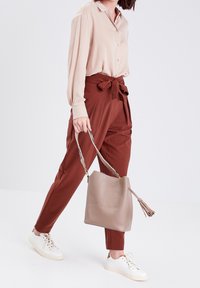 Chemise beige à manches longues, pantalon taille haute couleur rouille avec nœud, sac à main gris avec pompon, et baskets blanches, pose en marchant sur fond clair.
