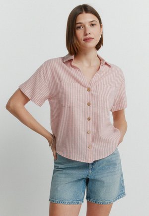 Frau mit kinnlangem braunem Haar, die ein pink-weiß gestreiftes kurzärmeliges Hemd mit Knöpfen und blaue Jeansshorts trägt und mit der Hand in der Hüfte posiert.