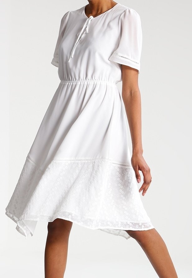 Valkoinen mekko, jossa on pyöreä pääntie, lyhyet pörröiset hihat ja joustava vyötärö. Helmassa pitsireunus, joka luo kerrosteisen vaikutelman.