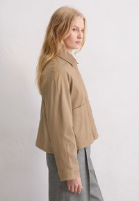 Beżowa kurtka z kołnierzem, przednimi kieszeniami i luźnym krojem. Materiał wydaje się gładki i wytrzymały, w połączeniu z szarymi spodniami.