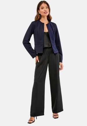 Marineblaues, cropped Jackett mit rundem Ausschnitt, glatter Textur und strukturierten Nähten, kombiniert mit schwarzen, weit geschnittenen Hosen und Riemchen-High Heels.