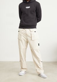 Uomo che indossa un cappuccio nero Calvin Klein, pantaloni cargo beige con molte tasche e scarpe slip-on bianche a scacchi, mentre sta in piedi su un pavimento beige.