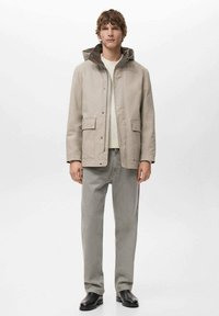 Młody mężczyzna w beżowej kurtce z kapturem, białym swetrze, szarych dżinsach i czarnych butach, stojący na tle jednolitego białego tła.