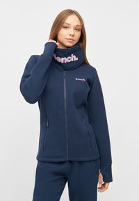 Námornícky modrý fleecový jacket na zips s vysokým golierom, s brand 'Bench.' v svetloružovej farbe na golieri a ľavej hrudi; hladká textúra.