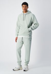 Felpa verde chiaro con cappuccio e cordini, tasche frontali e pantaloni jogger coordinati con tasche applicate e cintura elastica, abbinati a sneaker bianche.