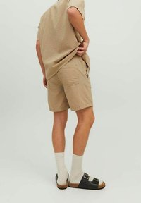 Personne portant un short et une chemise beige texturés, des chaussettes blanches et des sandales noires se tenant contre un fond clair uni.