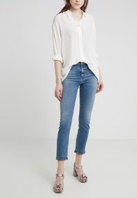 Camisa blanca de botonadura con mangas remangadas, combinada con unos vaqueros ajustados de color azul claro. Modelo llevando sandalias de tacón bloque con estampado. Atuendo simple y casual.