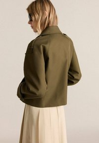 Olive green cropped jacket in smooth fabric, featuring a structured collar and prominent shoulder seams. Worn over a cream pleated skirt.