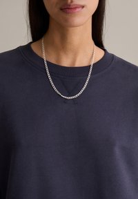 Srebrny naszyjnik z łańcuszkiem opada na granatową bawełnianą bluzę z okrągłym dekoltem i prążkowanymi detalami. Gładka faktura, minimalistyczne elementy.