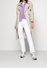 Gabardina beige con botones negros, llevada sobre una camisa de cuadros morados. Combinada con jeans rectos blancos y zapatillas deportivas grises.
