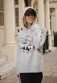 Gray knit hoodie with a large front graphic of cable cars and trees, featuring a textured hood and ribbed cuffs. Styled with navy pants.