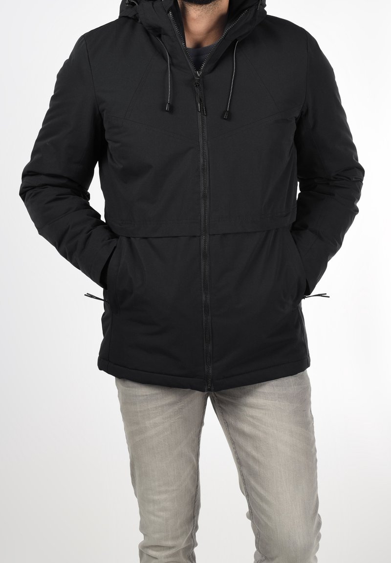 Homme portant une veste noire à capuche avec fermeture éclair, les mains dans les poches, et un jean gris clair sur un fond blanc uni.