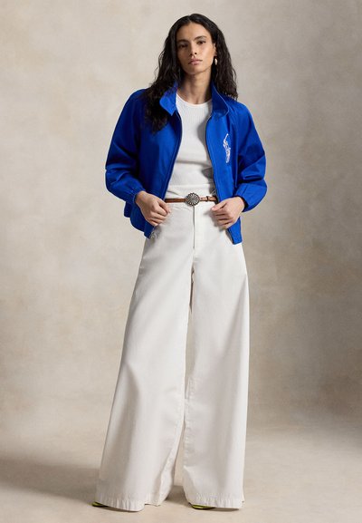 Vrouw die een blauw jack met ritssluiting draagt, een wit T-shirt, wijd uitlopende witte broek en een bruine riem met een decoratieve gesp, staande tegen een neutrale achtergrond.