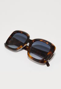 Gafas de sol con patrón de carey, con un marco cuadrado robusto, lentes tintados en azul y material de acetato. Presenta patillas gruesas.