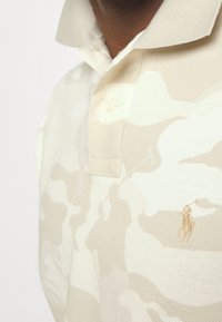 Beige polo tričko s camouflage vzorem, vyrobené z texturované tkaniny, s jemně vyšitým logem na hrudi a klasickým límcem.