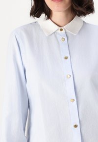 Donna che indossa una camicia azzurro chiaro con colletto bianco e bottoni dorati, accorciata all'altezza del mento e della parte superiore del torso.