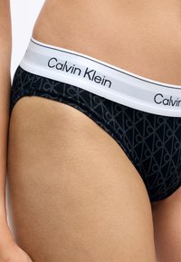 Czarne majtki bikini z geometrycznym wzorem, z białą elastyczną talią z napisem "Calvin Klein" w czarnych literach.