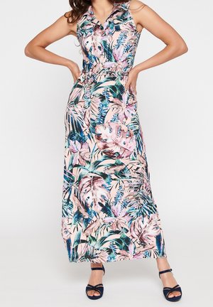 Kvinde iført ærmeløs, med krave, maxikjole med bælte med pink, grønne og blå tropiske bladmønstre og marineblå åbne hæle.