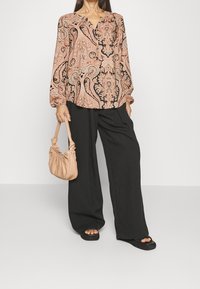 Langærmet bluse i paisley mønster i pink og sort, med V-hals og rynkede ærmer. Kombineret med brede sorte bukser og en blød beige taske.