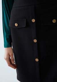 Černá sukně na knoflíky se zlatými detaily, s dvěma náprsními kapsami a hladkým povrchem. Zelená halenka s dlouhým rukávem je částečně viditelná.