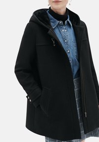 Manteau en mélange de laine noir avec une capuche, fermeture éclair, poches latérales et poignets à boutons, porté sur une chemise en denim bleu et une jupe à carreaux.