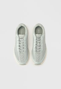 Αθλητικά παπούτσια ανοιχτού γκρι χρώματος με υφασμάτινη επάνω επιφάνεια με υφή, στρογγυλεμένη μύτη, λευκή αντιθετική σόλα και κλείσιμο με κορδόνια. Το λογότυπο "Lacoste" είναι ορατό στη γλώσσα του παπουτσιού.